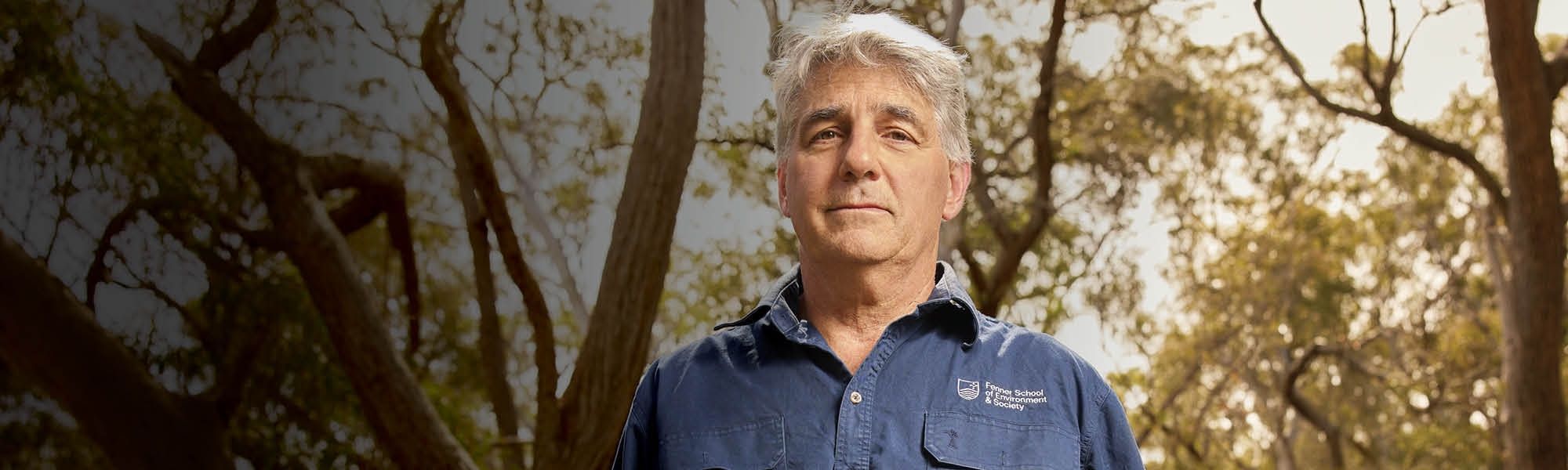 an older man standing confidently outdoors in front of tall trees