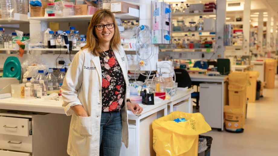 Dr Vicki Athanasopoulos is in a science lab wearing a white lab coat. She is wearing a floral pattern shirt under her lab coat